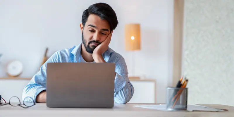 A Man Sitting In Front Of His Laptop Looking Bored During A Video Call, Showing Lack Of Engagement And Poor Camera Presence.