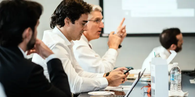 Business Professionals In A Meeting, One Checking His Phone While Others Talk — Highlighting Distractions During Video Calls.